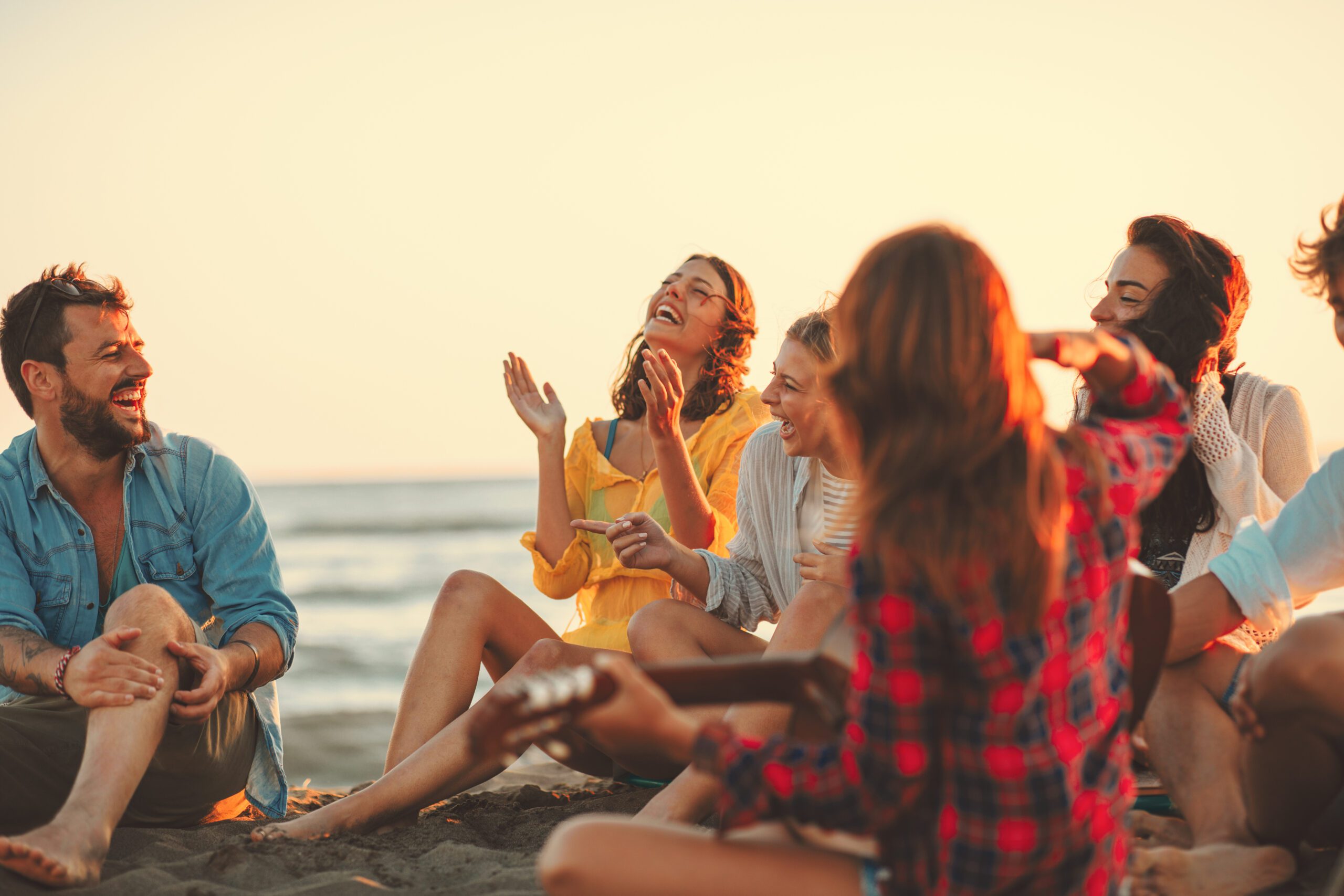 Some friends sit on a beach with the sun setting behind then. Some smile and laugh while others play the guitar.