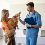 A woman in an orange shirt is holding a corgi and talking to a vet wearing blue scrubs. The vet is petting the dog.