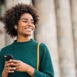 A smiling woman wearing white Bluetooth earbuds and a sweater walks outside holding her mobile phone in her hands.