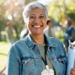A smiling woman in a denim jacket standing on a park path, wearing a lanyard and holding a clipboard with papers.