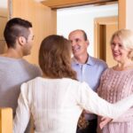 A man and woman greet an older man and woman in an open doorway. All four people are smiling.