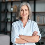 A confident, stylish middle-aged woman with gray hair smiles and poses with her arms crossed in her home office.