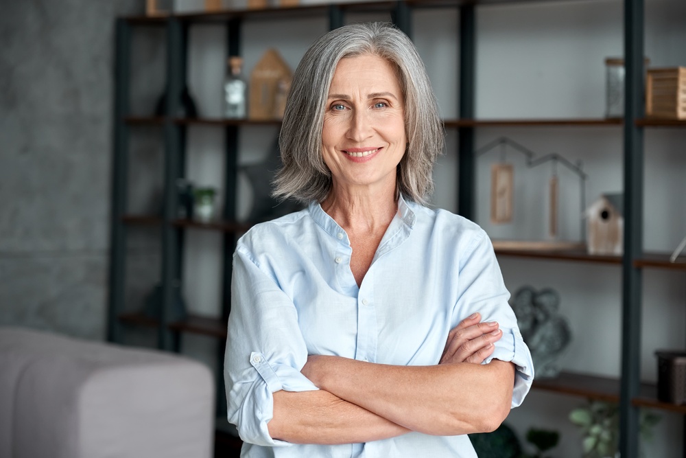 A confident, stylish middle-aged woman with gray hair smiles and poses with her arms crossed in her home office.