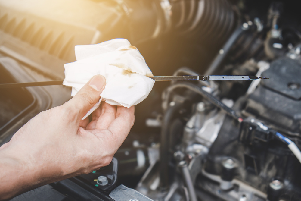 A close-up view shows a hand gripping a white cloth around an oil dipstick from a vehicle. The engine is behind the stick.