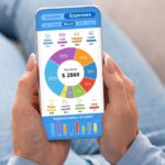 Home 9 A person holds a mobile phone in their hands. The phone screen displays a pie chart of monthly expenses.