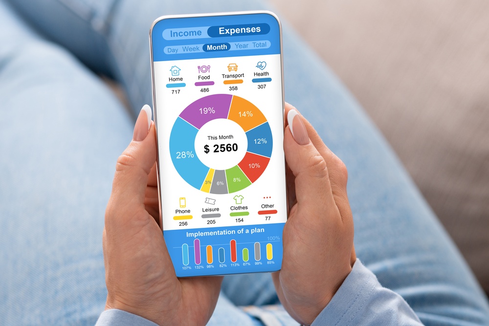 A person holds a mobile phone in their hands. The phone screen displays a pie chart of monthly expenses.