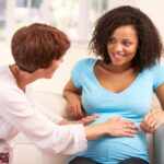 A happy, Black pregnant woman in a blue shirt sitting down as the midwife presses her abdomen for examination.