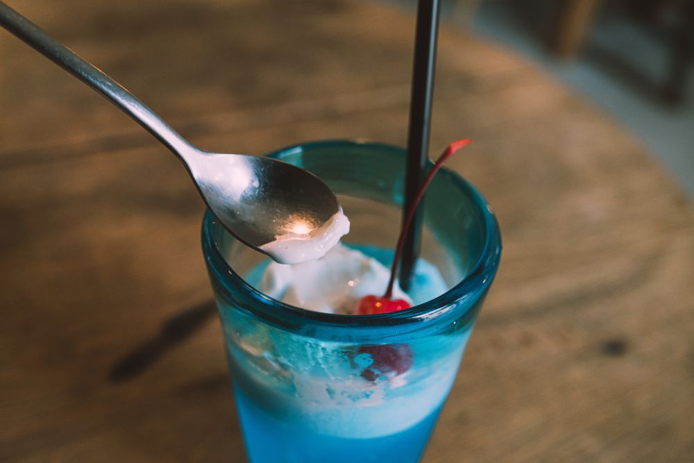 A glass with blue soda, ice cream, a cherry, and a straw sticking out of it while a spoon scoops some ice cream.