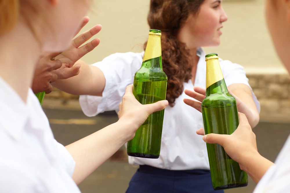 A young girl in a school uniform turns her head away and pushes back two beers offered to her by her peers.