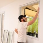 A young man in a black cap examines a window frame, with new windows leaning against the wall beside him.