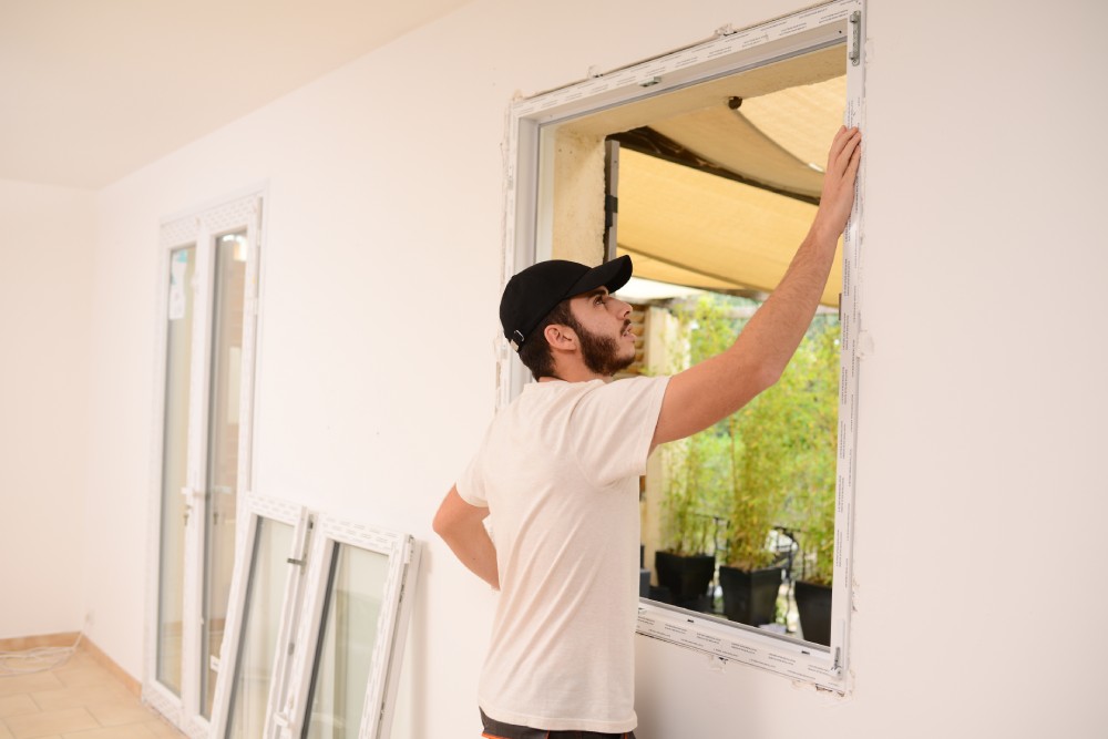 A young man in a black cap examines a window frame, with new windows leaning against the wall beside him.