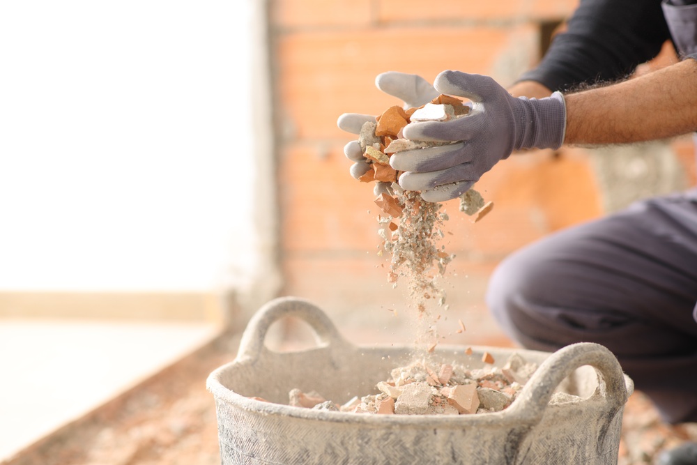 A close-up of a man with gloves dropping demolished stone and concrete into a large basket full of debris.