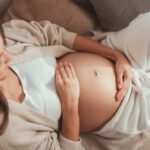 An overhead view of a pregnant woman lounging on a tan couch, placing both hands on her belly.