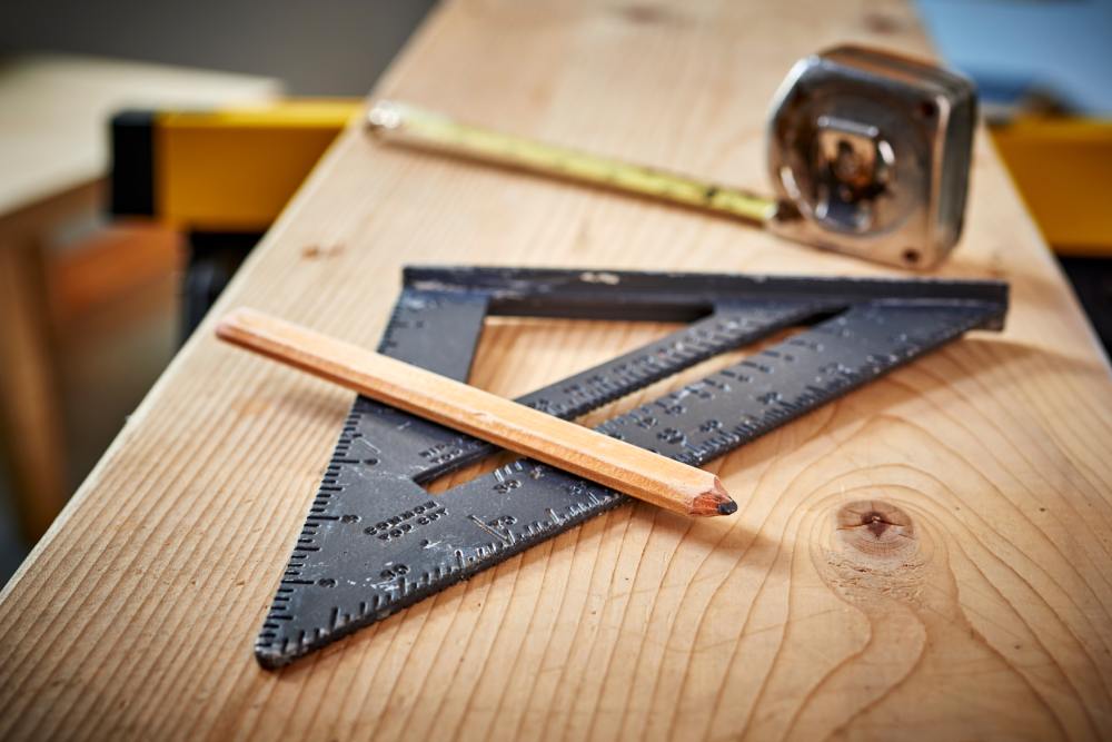 A piece of wooden plank with a pencil, a tape measure, and a worn-out black set square sitting on top of it.