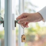 A person's hand inserting keys into the doorlock of a home. A wooden cutout of a house dangles from the key ring.