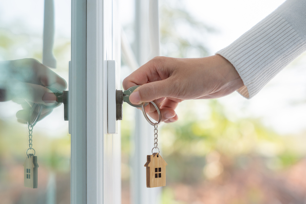A person's hand inserting keys into the doorlock of a home. A wooden cutout of a house dangles from the key ring.