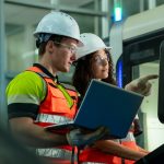 A man and a woman wearing white hard hats, protective goggles, and hi-vis vests use a computer. The man points to the screen.
