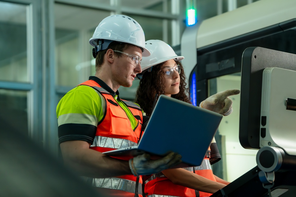 A man and a woman wearing white hard hats, protective goggles, and hi-vis vests use a computer. The man points to the screen.