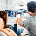 A dentist holds up a patient's X-rays to explain a problematic tooth to the patient, who asks him a question.