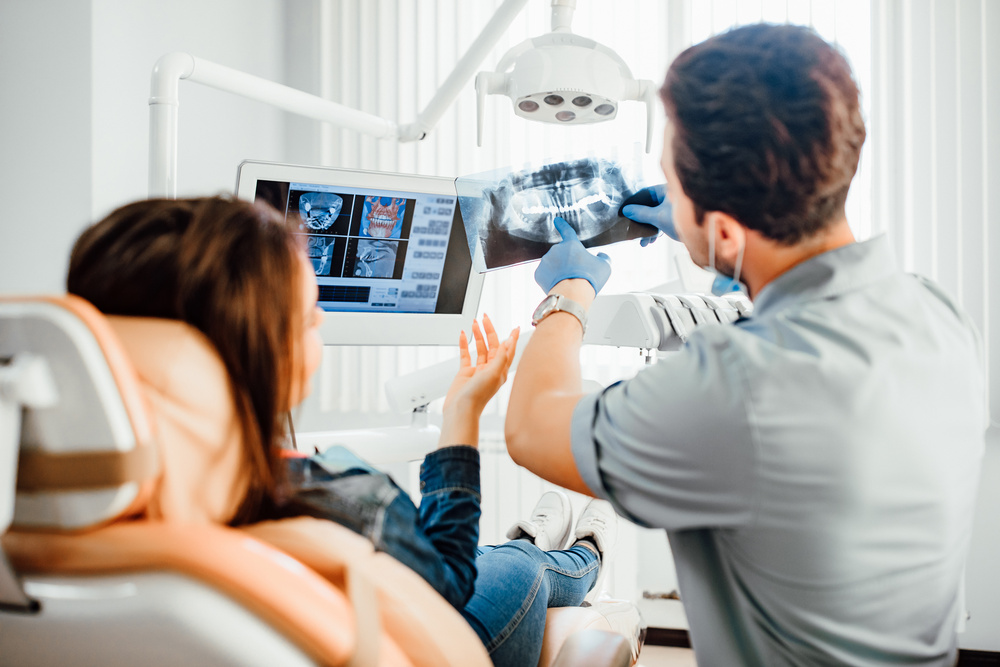 A dentist holds up a patient's X-rays to explain a problematic tooth to the patient, who asks him a question.
