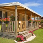 A gray mobile home with charcoal shutters features a wooden terrace with red hanging flowers and pink blooms below.