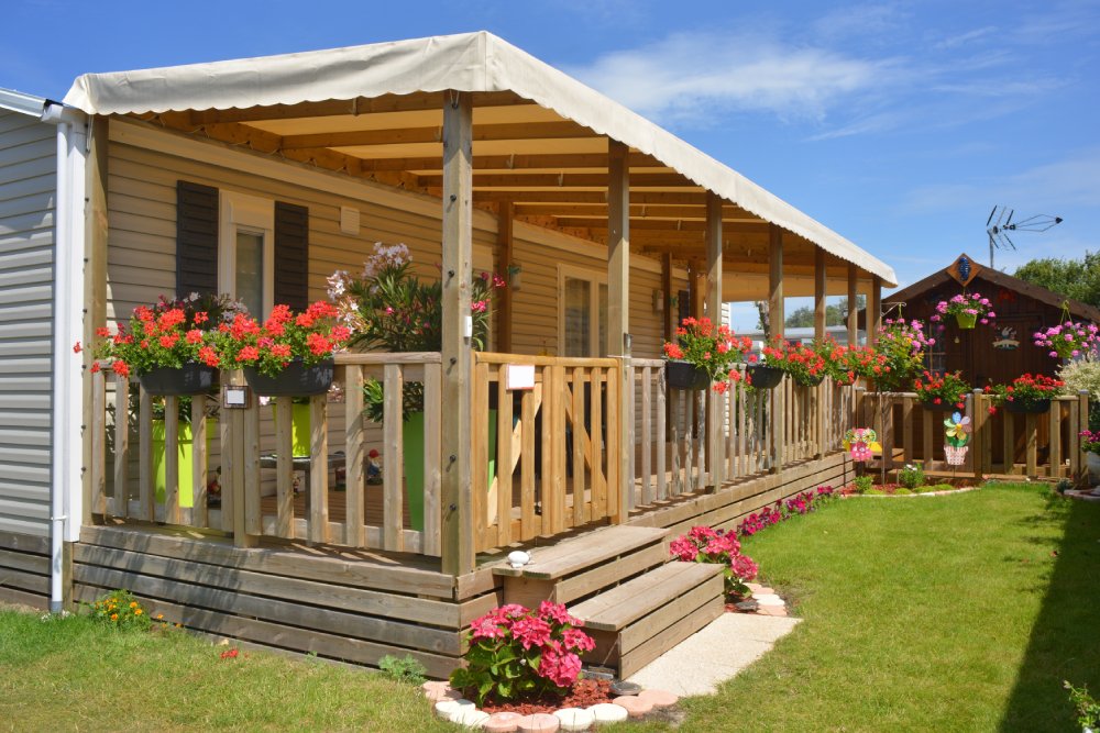 A gray mobile home with charcoal shutters features a wooden terrace with red hanging flowers and pink blooms below.