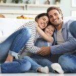 A smiling and happy young man and woman sit on the floor, embracing a happy little girl. A white couch is behind them.