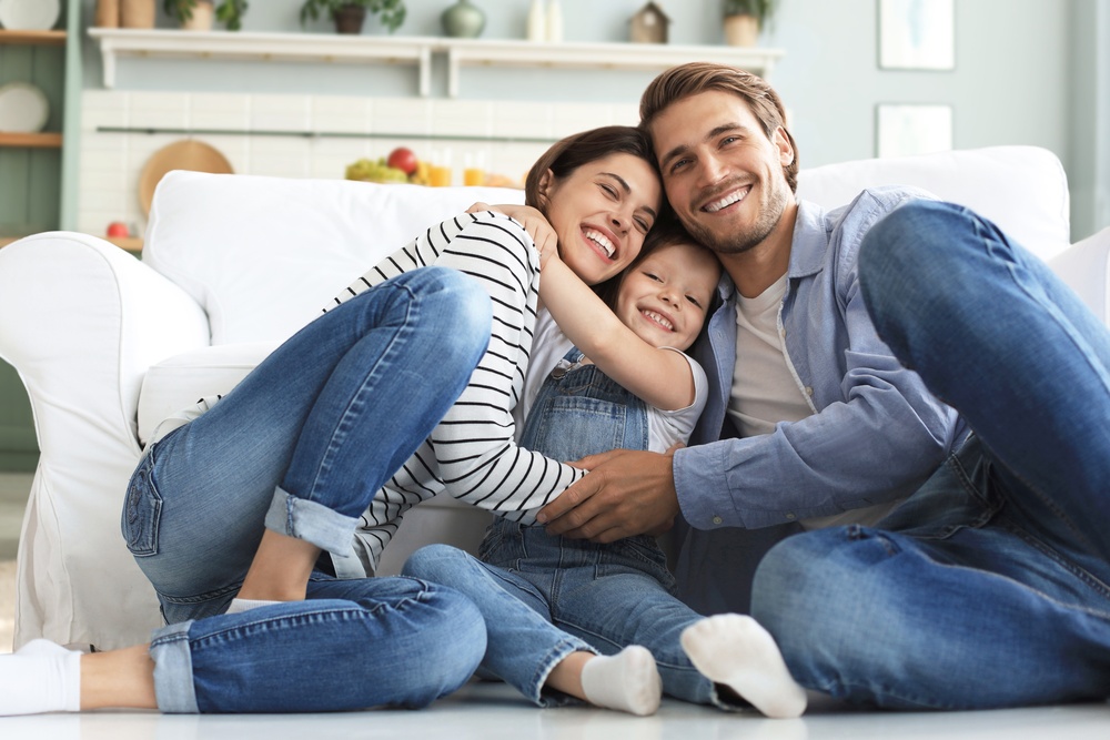 A smiling and happy young man and woman sit on the floor, embracing a happy little girl. A white couch is behind them.