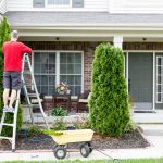 A man in a red shirt is standing on a ladder working on his home's roof and drainage system in front of his landscaping.