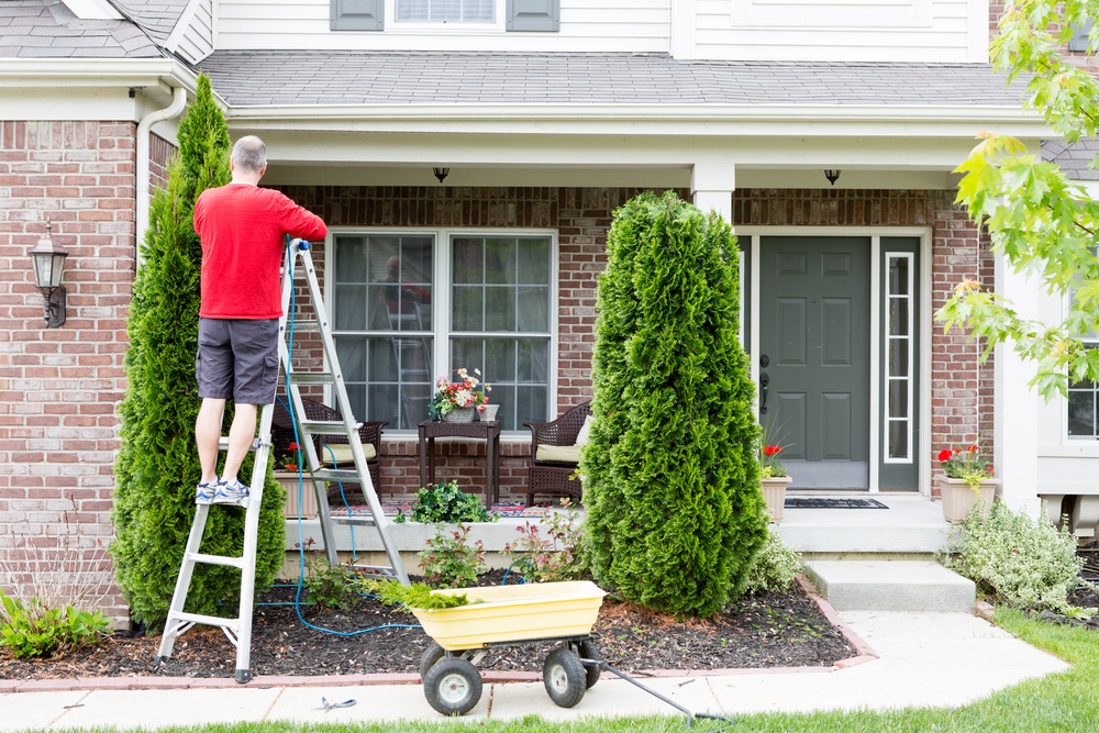 A man in a red shirt is standing on a ladder working on his home's roof and drainage system in front of his landscaping.