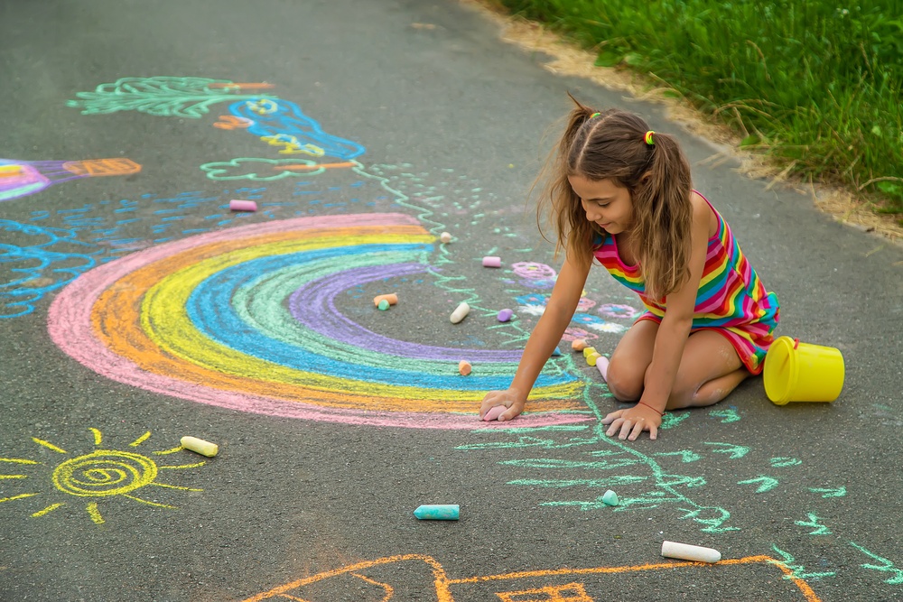 A small child with their hair up kneeling onto asphalt and using various sticks of colorful chalk to draw pictures.
