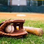 A brown baseball glove with a baseball inside and a wooden bat sitting on the grassy area of a baseball field.