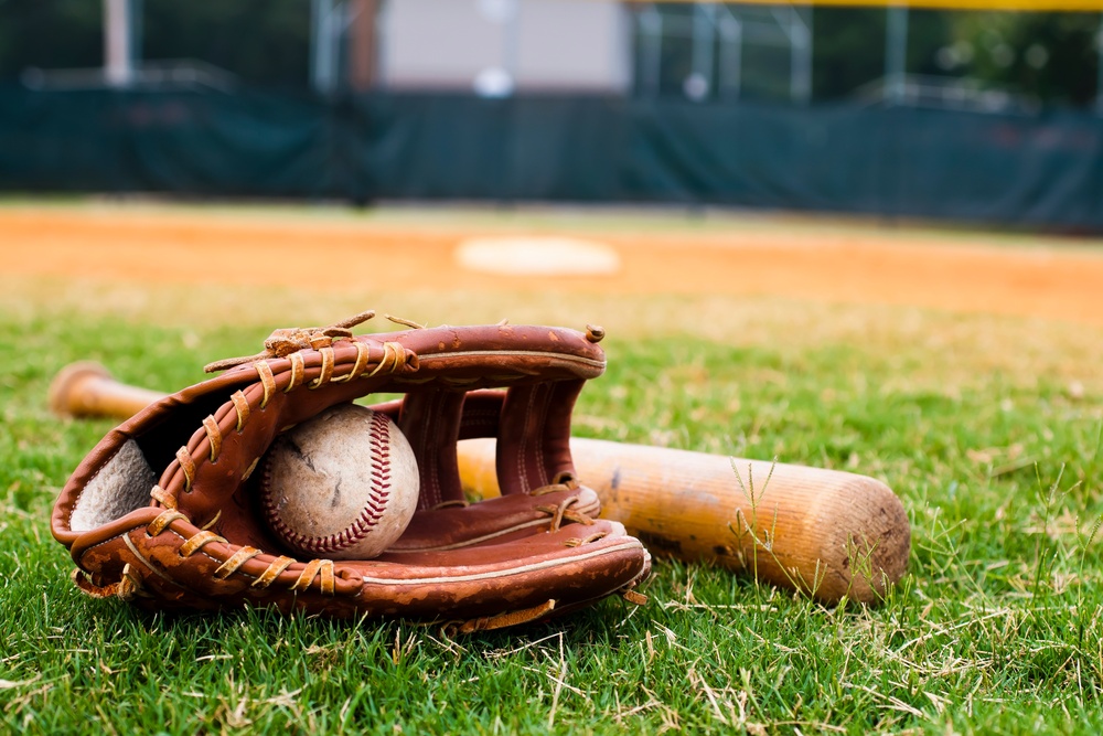 A brown baseball glove with a baseball inside and a wooden bat sitting on the grassy area of a baseball field.