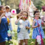 Five children blowing bubbles at an outdoor party. Colorful decorations are dangling above the children.