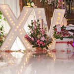 Three arrangements of pink and white flowers sit in front of a large illuminated "X" and “V” that have light bulbs in them.