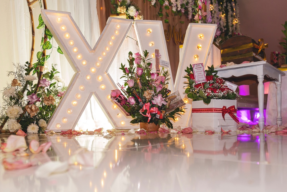 Home 2 Three arrangements of pink and white flowers sit in front of a large illuminated "X" and “V” that have light bulbs in them.