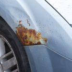 The front of a car that has some rusting by the wheel well. The car is blue with silver tire rims.