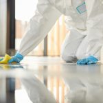 A person is kneeling on the ground and cleaning up a mess. They are wearing blue gloves and a white jumpsuit.