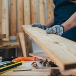A close-up view shows a person wearing gray protective gloves while moving a piece of plywood in a woodshop.