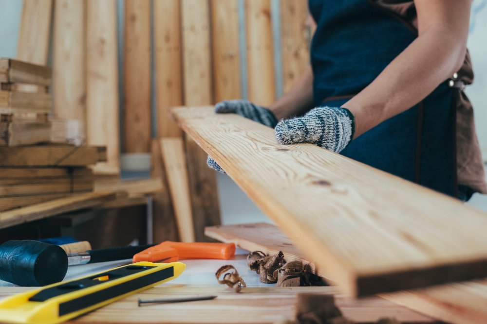 A close-up view shows a person wearing gray protective gloves while moving a piece of plywood in a woodshop.