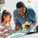 A father shows his daughter how to check the oil in the family car. He wipes the dipstick with a yellow rag.