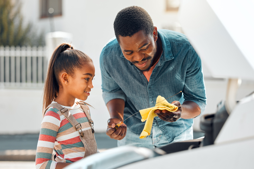 A father shows his daughter how to check the oil in the family car. He wipes the dipstick with a yellow rag.