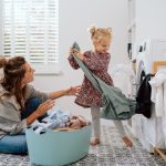 A woman sitting with a laundry basket in a laundry room while a young girl pulls a sweatshirt from the dryer.