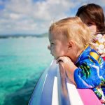Two children in Hawaiian shirts leaning over the side of a boat, gazing into the ocean beneath a partly cloudy sky.