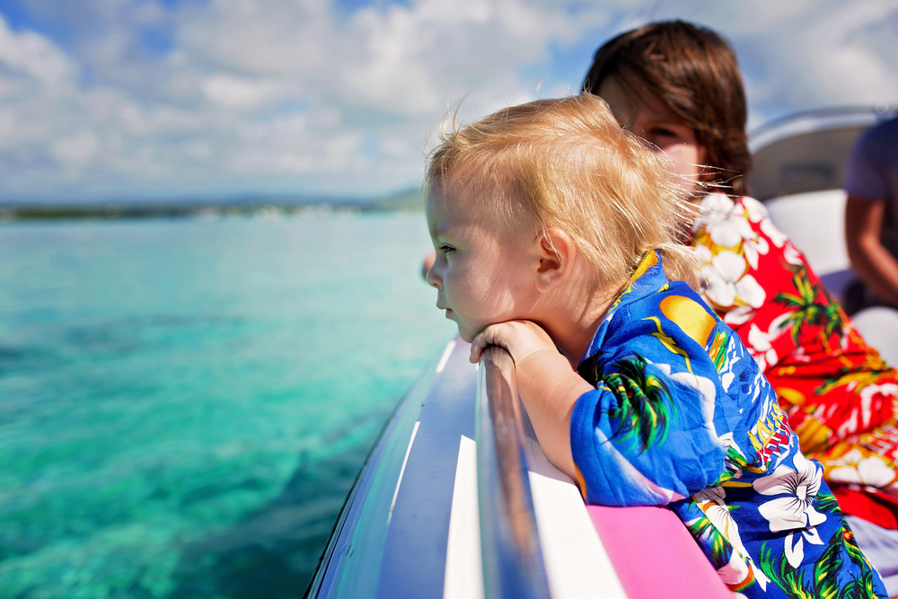 Two children in Hawaiian shirts leaning over the side of a boat, gazing into the ocean beneath a partly cloudy sky.