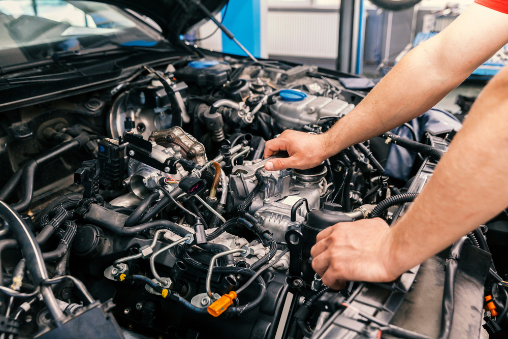 A close-up view shows a person leaning on the front of a vehicle that has its hood popped open, exposing the engine.