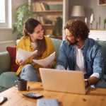 A happy couple sits together on their couch reviewing important documents while a laptop is open on the coffee table.