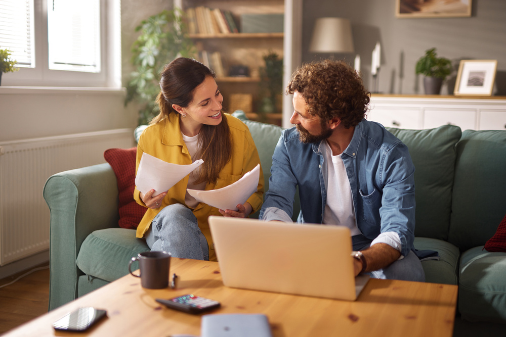 A happy couple sits together on their couch reviewing important documents while a laptop is open on the coffee table.