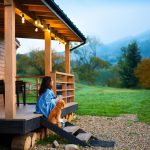 A woman wrapped in a blue blanket holds a blue mug while sitting on the front steps of a cabin with her dog beside her.