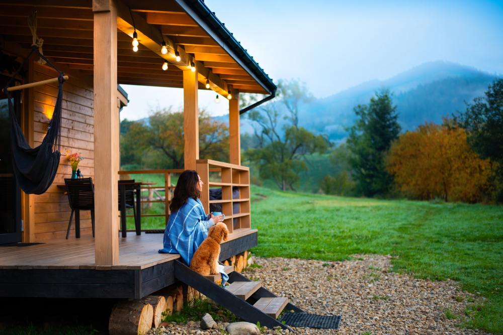 A woman wrapped in a blue blanket holds a blue mug while sitting on the front steps of a cabin with her dog beside her.
